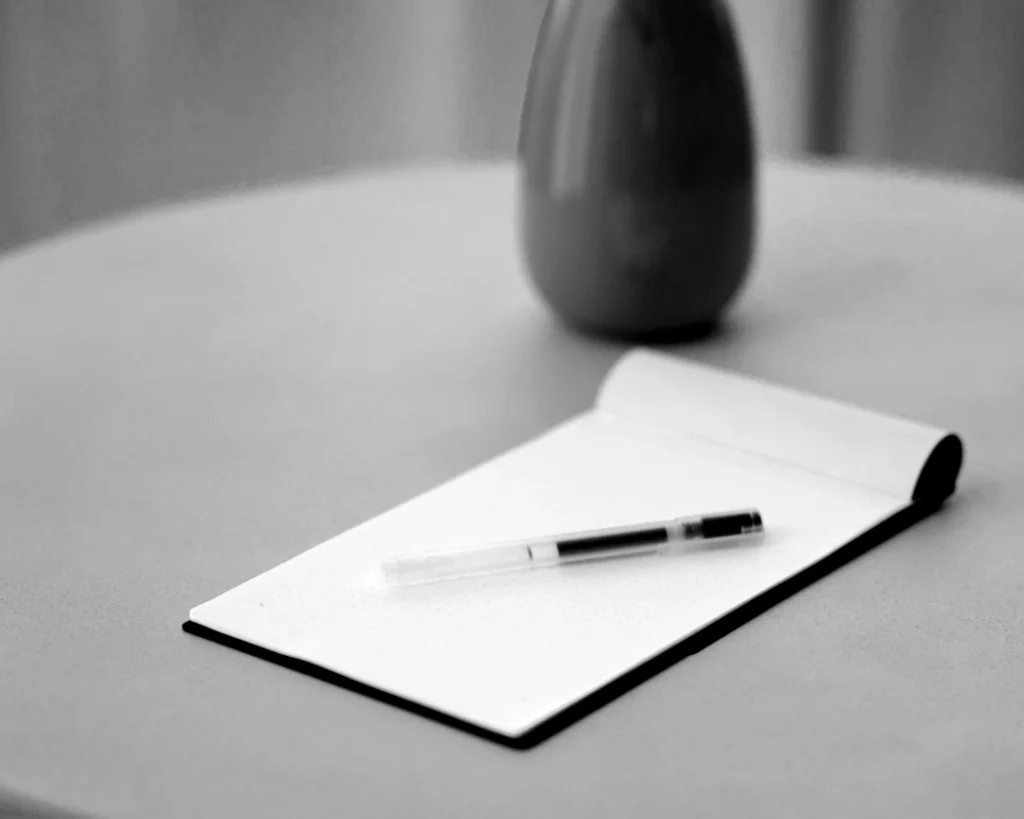 Schwarz-weißes Bild eines aufgeschlagenen, leeren Notizbuchs mit einem Stift auf einem runden Tisch, passend zu den Themen Masterclasses, Fokus und Corporate Wellbeing.
