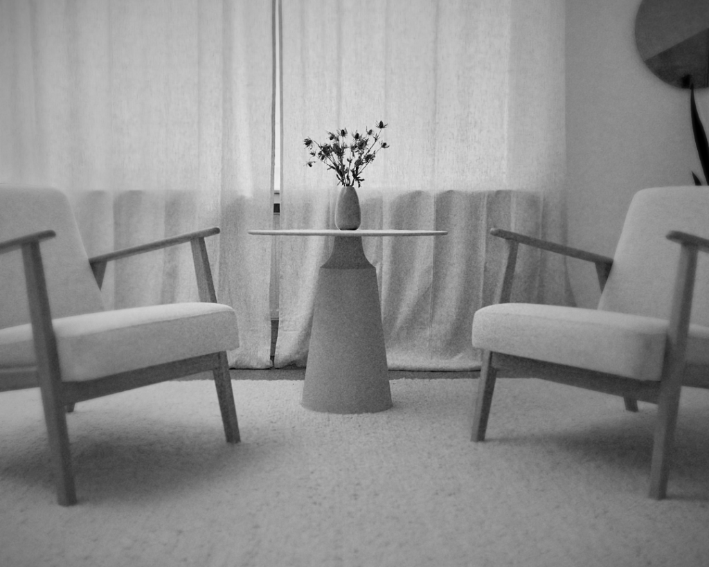 Schwarz-weißes Foto des einladenden Behandlungsraums in The Sanctuary: zwei bequeme Sessel mit Holzrahmen, ein moderner konischer Beistelltisch mit einer Vase getrockneter Blumen, vor einem Fenster mit leichten Vorhängen. Ein Raum, der Privatsphäre und Ruhe ausstrahlt.